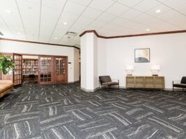 A spacious waiting area with two armchairs, a console table with lamps, and a view through glass-paneled double doors into a library with bookshelves.
