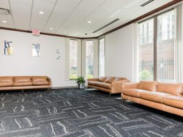 A waiting room with three tan leather sofas, dark patterned carpeting, and framed wall art.
