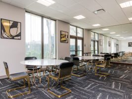 A modern lounge area with two round tables, each with four black chairs featuring gold-toned metal bases, set against a wall with framed abstract art and a large mirror.