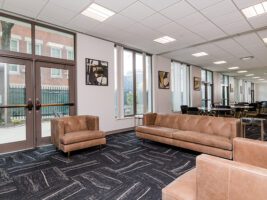 A waiting area with leather sofas, armchairs, and a conference table, featuring large windows and patterned carpet.