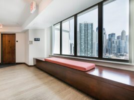 A hallway with a wooden built-in bench featuring a red cushion, set against a large window wall overlooking a city skyline.