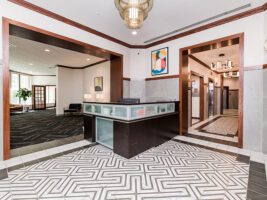 A hotel or office lobby featuring a central reception desk, geometric floor tiling, and dark wood trim around arched entryways leading to adjacent rooms and elevators.