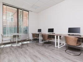 A bright office room with a small round table and chairs by a window, and a row of three computer workstations along a white wall.