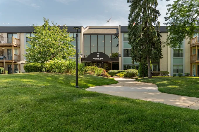 The exterior of a multi-story apartment building with a Greenleaf Apartments sign above the main entrance, situated behind a grassy lawn and a curved walkway.