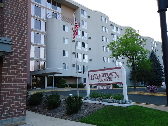 Exterior of Rivertown Commons apartment building with a sign in the foreground and an American flag flying near the entrance.